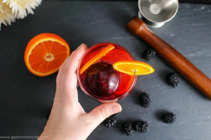 Blackberry Tangerine Vodka Tonic Cocktail in glass, hand holding it. Orange half, blackberries, muddler and jigger