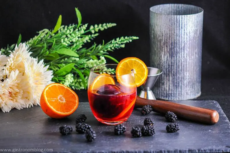 Blackberry Tangerine Vodka Tonic Cocktail in glass with orange half and wheel. Blackberries, muddler, jigger. Flowers and metal container in background
