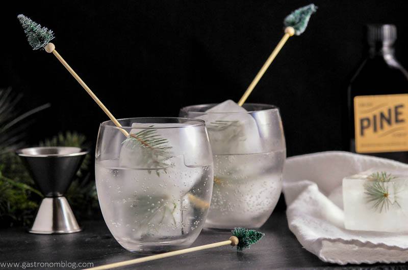 Mountain Pine Gin and Tonic with pine infused ice cubes and pine tree topped cocktail stirrers. Jigger and Dram Apothecary Syrup bottle in background