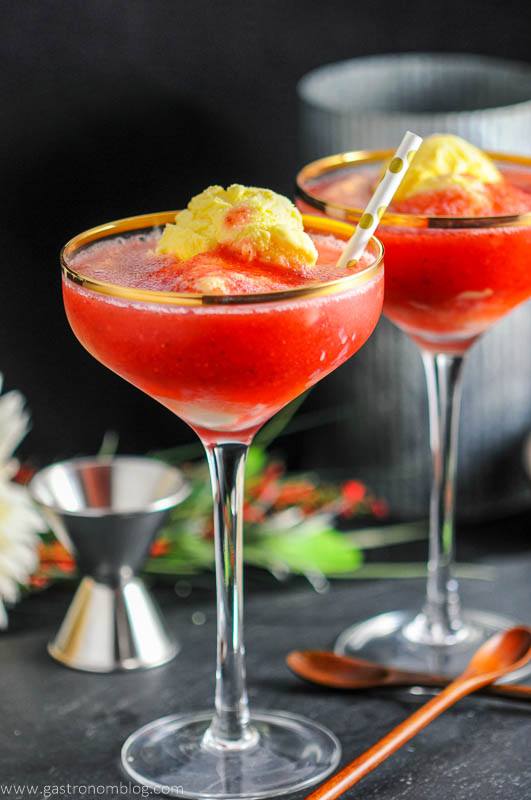 Strawberry Rhubarb Daiquiri Float, red cocktails with ice cream in gold rimmed coupes with gold dot straws. Jigger, wood spoon and flowers in background