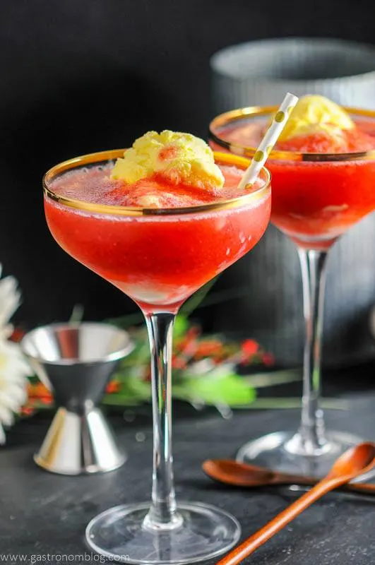 Strawberry Rhubarb Daiquiri Float, red cocktails with ice cream in gold rimmed coupes with gold dot straws. Jigger, wood spoon and flowers in background
