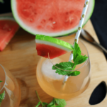 Top shot pink cocktail with mint, watermlon slice on cutting board, half a watermelon