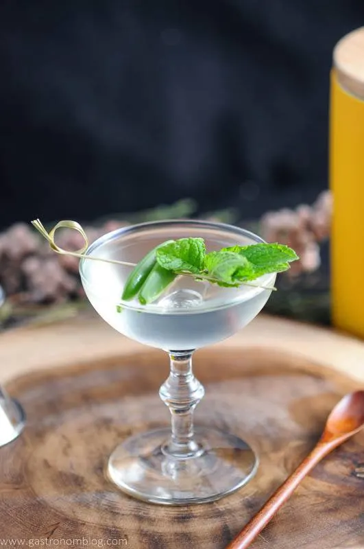 The Spring is Here cocktail with mint and peas in coupe on wooden plate. Wooden spoon, flowers and yellow canister in background