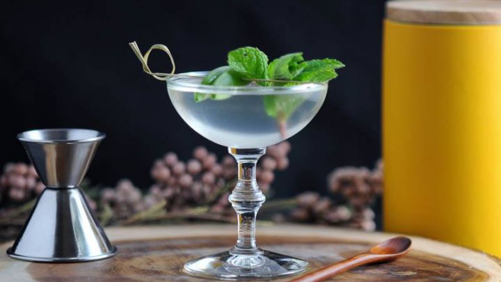 The Spring is Here, clear cocktail in coupe with mint and pea on cocktail pick. Yellow canister, jigger, wooden spoon and flowers in background