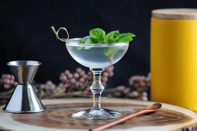 The Spring is Here cocktail in a coupe with mint and peas. Jigger, woodpen spoon, flowers and yellow canister in background