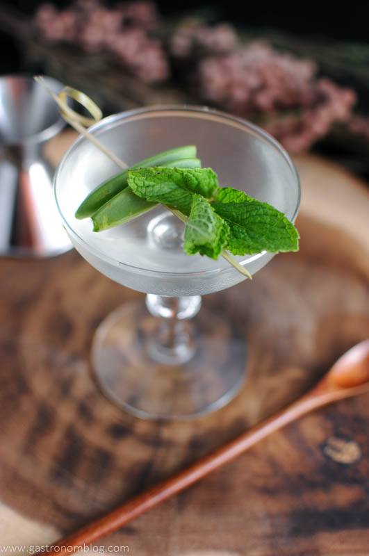 The Spring is Here, clear cocktail in coupe with pea and mint on cocktail pick. Wooden spoon and flowers