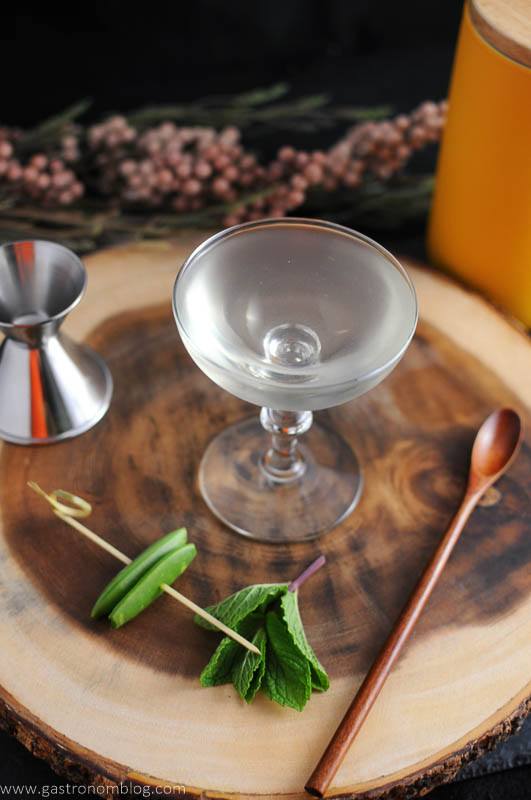 The Spring is Here cocktail in coupe. Mint and peas with jigger and wooden spoon on wooden plate. Flowers and yellow canister in background