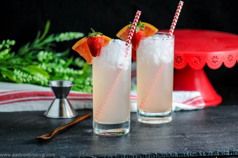 Strawberry Paloma cocktail in highballs with red straws, strawberry and grapefruit wedge. Wood Spoon, jigger, red cake stand and flowers in background