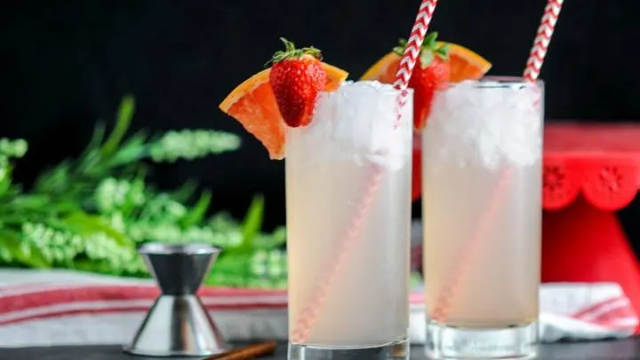 Strawberry Paloma, pink cocktails in highballs, red straws, strawberries and grapefruit slices. Greenery, jigger and red stand in background