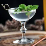 The Spring is Here Cocktail in a coupe with mint and fresh peas as garnish on a wood board from Gastronomblog