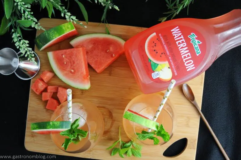 cocktail top shot. 2 glasses with watermelon wedges, mint and straws. On cutting board with watermelon slices, jigger, wooden spoon and jigger. Flowers in background