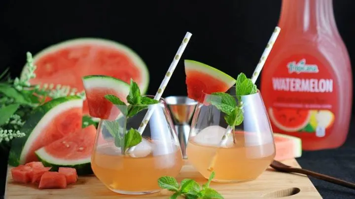 Watermelon Bourbon Cooler, pink cocktails in glasses with mint, watermlon slices and dot straws. Watermelon, mint and watermelon bottles in background