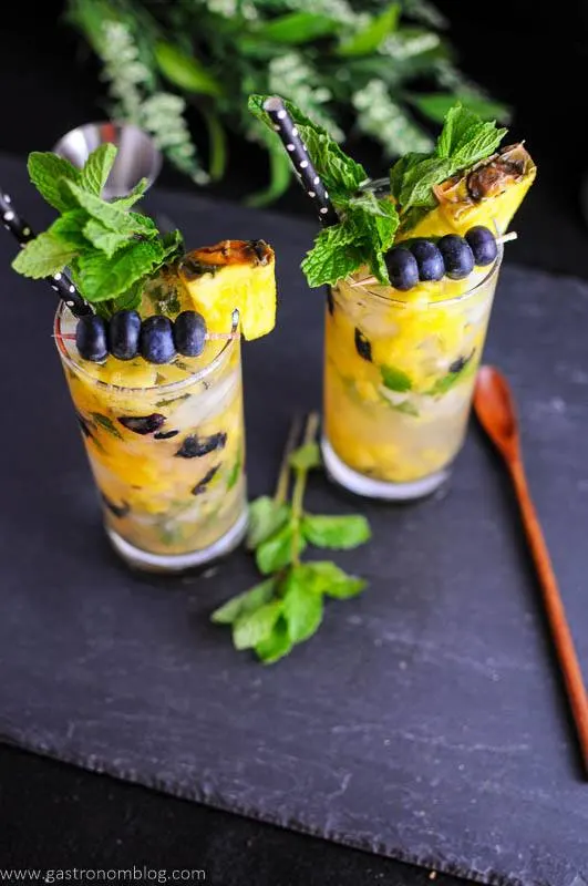 Highball glasses filled with pineapple and blueberries, mint on top. Wooden spoon on slate.