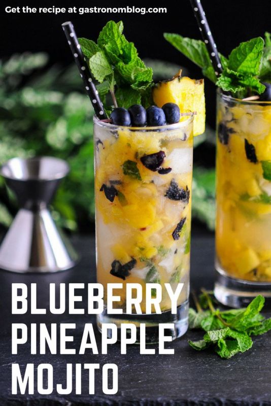 Blueberry Pineapple Mojito, 2 glasses filled with pineapple and blueberries. Pineapple, blueberries and mint on top with black and white dot straws. Jigger and greenery in background