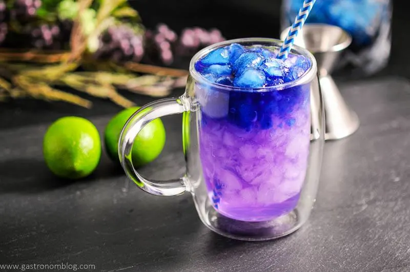 Galaxy Magic Moscow Mule in clear glass mug with blue straw. Limes, flowers and jigger in background. Color changing cocktail