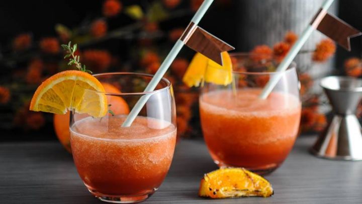 Tangerine Frozen Negroni, orange cocktails in glasses with orange slices and straws. Metal container, orange flowers in background