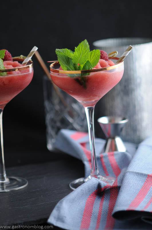 Watermelon Raspberry Frosé in cocktail coupes, mint and straws. Blue napkin, jigger and metal canister in background. 