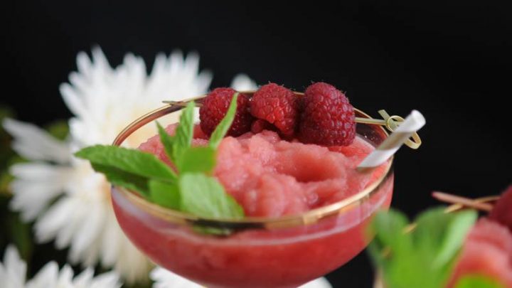Watermelon Raspberry Frosé, pink cocktail in gold rimmed coupe with raspberries and mint. White flowers behind