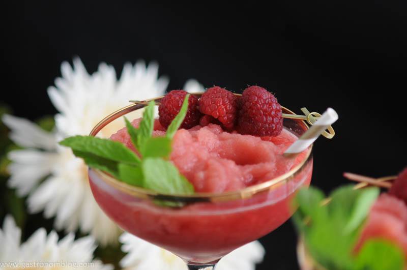 Watermelon Raspberry Frosé in gold rimmed cocktail coupe. With Straw, raspberries and mint. Flowers in background. 