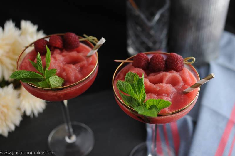 Watermelon Raspberry Frosé in cocktail coupes. Straws, raspberries and mint. Flowers, blue napkin in background. 
