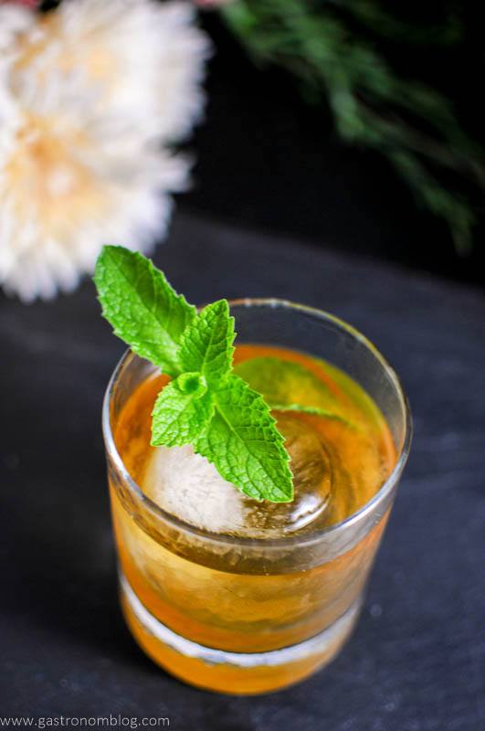 Apricot Honey Bourbon Sour Cocktail, yellow cocktail in rocks glass with ice ball and mint, white flowers behind