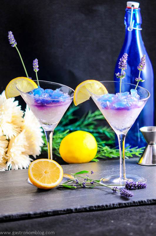 Lavender Lemon Gin and Tonic Granita Cocktails in martini glasses with lemon slices and lavender leaves. Lemons, blue bottle, jigger and flowers in background