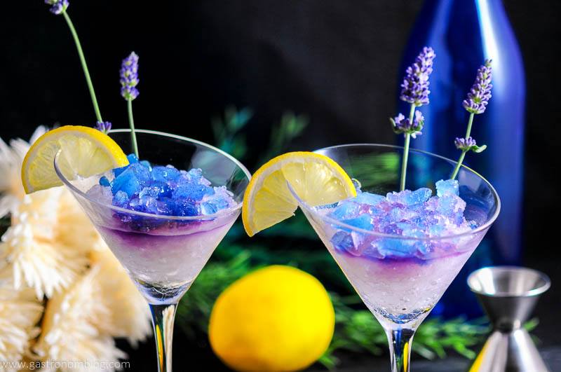 Lavender Lemon Gin and Tonic Granita Cocktail in martini glasses with lemon slices and lavender flowers. Blue bottle, flowers and lemon with jigger in backgrond