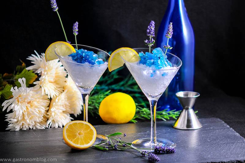 Lavender Lemon Gin and Tonic Granita Cocktails in martini glasses with lemon slices and lavender flowers. Flowers, blue bottle, jigger and lemon in background
