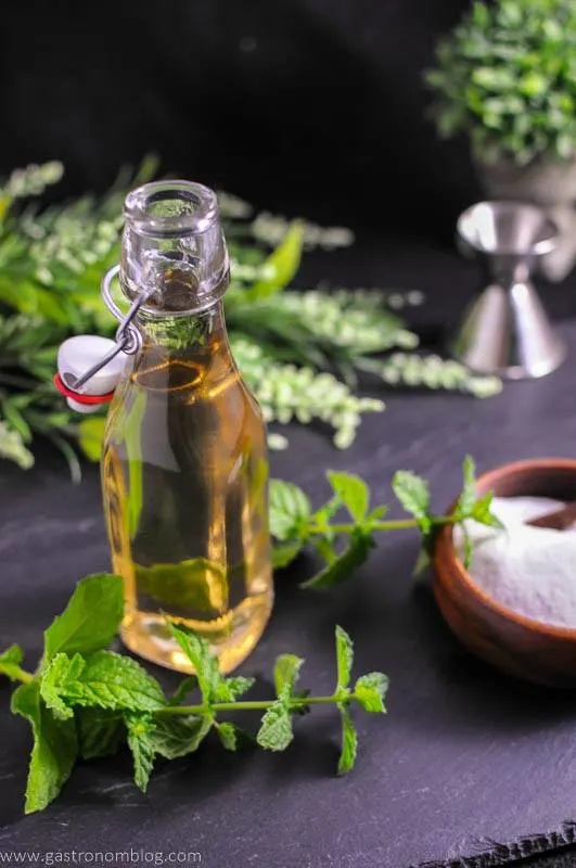Mint Simple Syrup - green syrup in bottle with mint leaves around, Wooden bowl of sugar, silver jigger and greenery in background