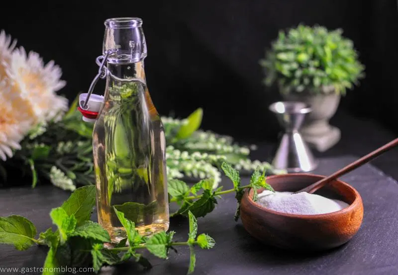 Mint Simple Syrup in bottle with wooden bowl filled with sugar. Mint leaves, flowers and jigger in background
