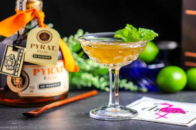 The Shipwreck cocktail in coupe with mint. Rum bottle, flowers, wood spoons, flamingo napkin in background