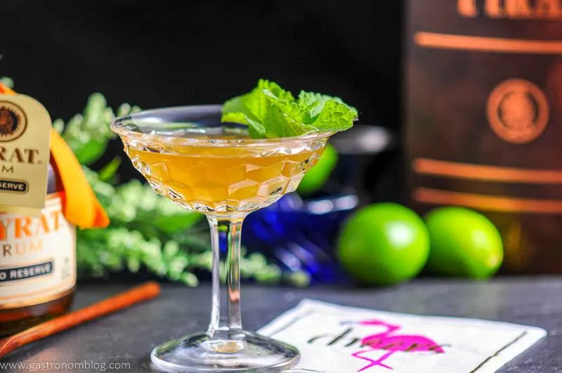 The Shipwreck cocktail in coupe with mint. Rum bottle, wood spoon, flamingo napkin and limes in background