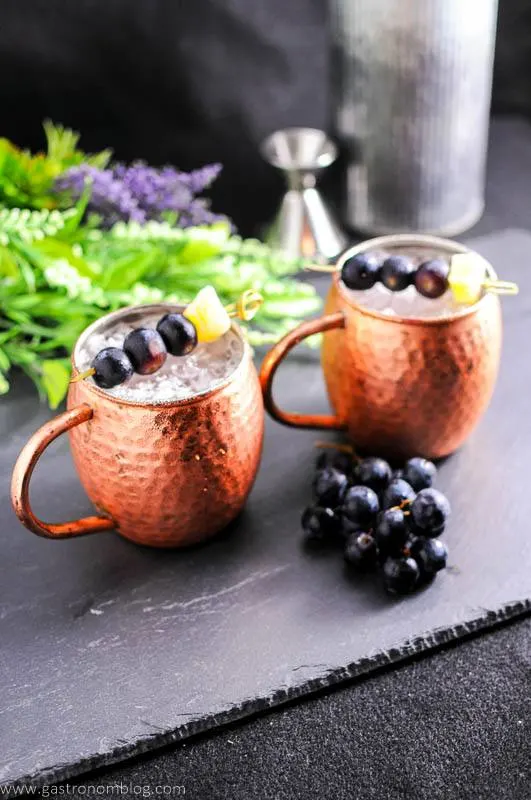 Concord Grape version of the classic Moscow Mule. Ginger beer, vodka, lime juice and Concord Grape simple syrup from Gastronomblog. #cocktail #lime #grapes #recipes #gastronomblog Concord Grape Moscow Mule in copper mugs with grapes and ginger on cocktail pics. Bunch grapes, flowers, jigger and metal canister in background