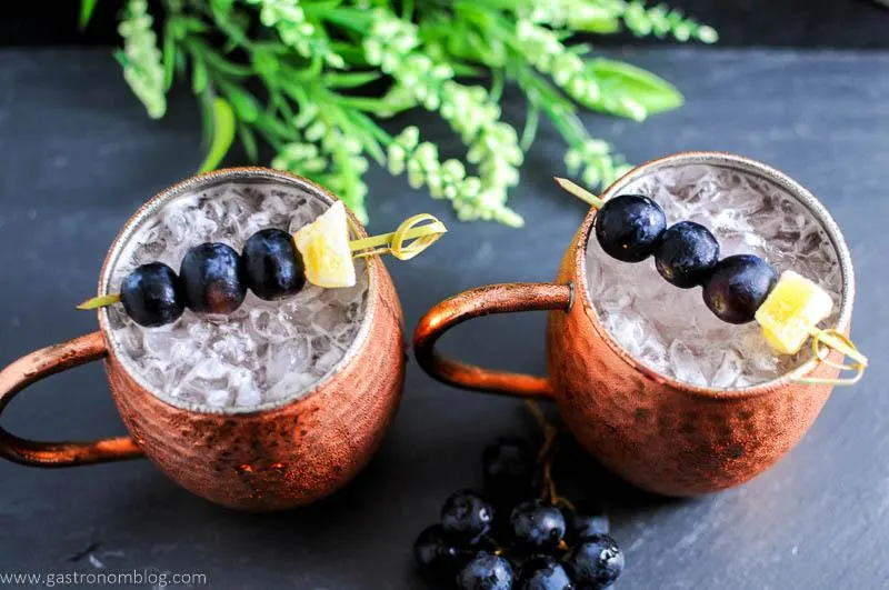 Concord Grape Moscow Mule in copper mugs. Grapes and ginger on cocktail picks. Flowers in background