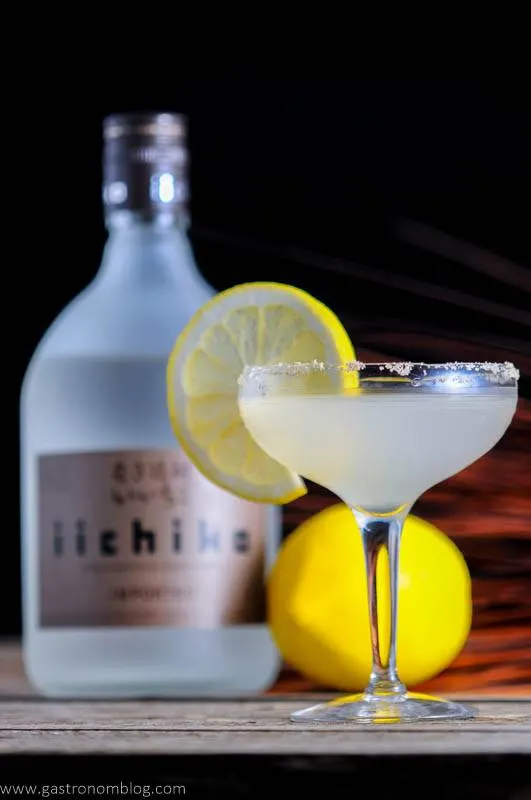 The Japanese Sidecar in cocktail coupe with lemon wheel. Lemon and iichiko bottle in background