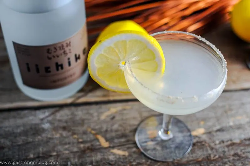 The Japanese Sidecar in cocktail coupe with lemon wheel. Lemons and iichiko bottle in background