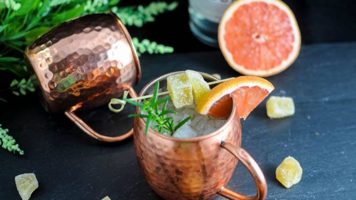 Rosemary Grapefruit Moscow Mule, in copper mugs with candied ginger, rosemary and grapefruit slices. Vodka bottle and flowers in background