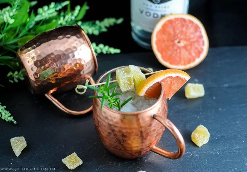 Rosemary Grapefruit Moscow Mule in copper mugs with grapefruit slices, ginger and rosemary. Vodka bottle and flowers in background