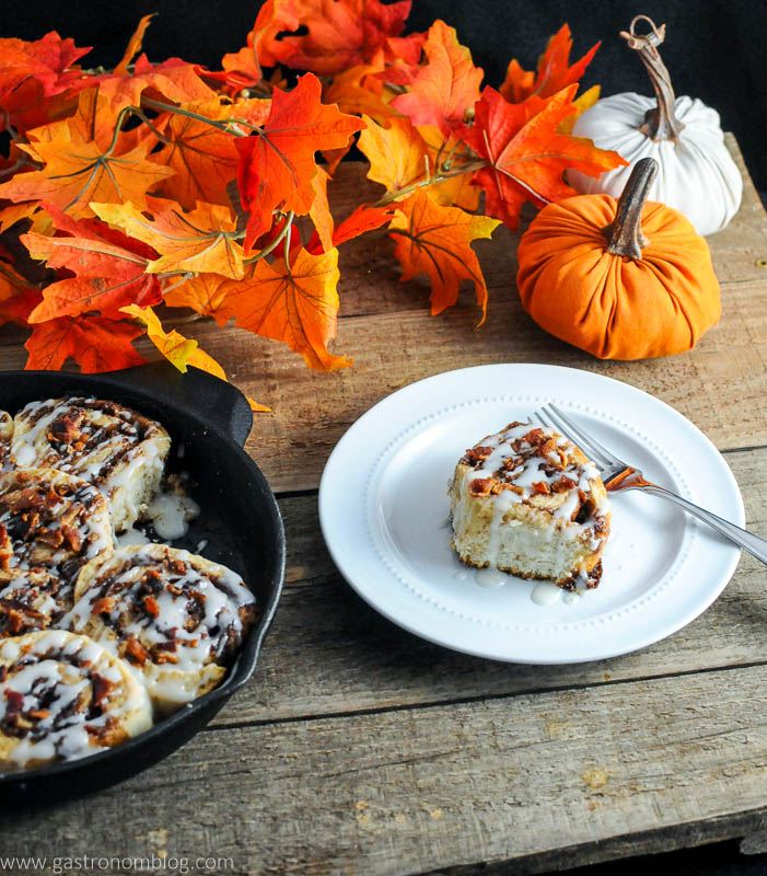 Bourbon bacon cinnamon rolls topped with bacon pieces on white plate with fork. Pumpkin and fall leaves with rolls in skillet.