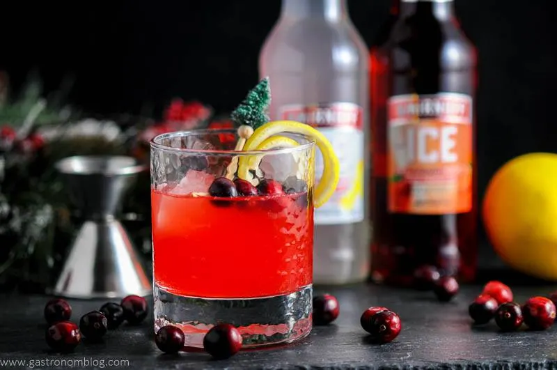 Holiday Berry Sparkler Cocktail in rocks glass with cranberries, lemon peel and pine tree topped stirrer. Jigger, cranberries, lemon and Smirnoff Ice bottles in background
