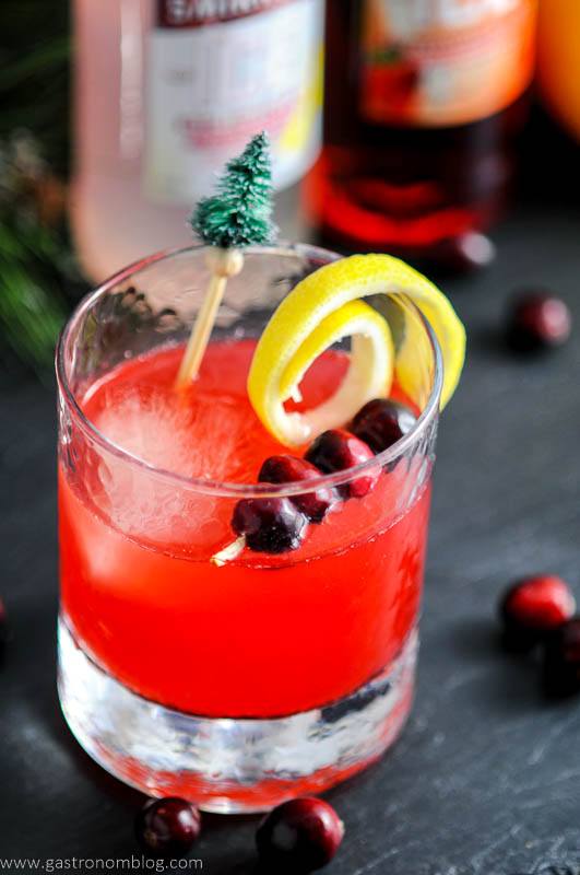 Cranberries and lemon curls garnish our Holiday Berry Sparkler Cocktail in a rocks glass, pine tree topped cocktail stirrer. Smirnoff bottles in background