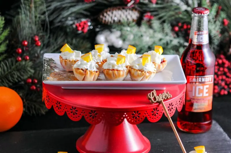 These Mini Berry Bites are the perfect Holiday party dessert. Easy to eat, and serve. These boozy strawberry gelatin filled cups are topped with whipped cream and oranges from Gastronomblog. #dessert #gastronomblog #strawberry #holiday #bitesize Mini Berry Bites with Smirnoff Ice Bottle and red cake plate