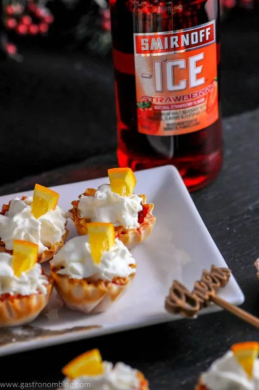 Mini berry bites in phyllos shells, topped with whipped cream on white plate. Red beer bottle behind