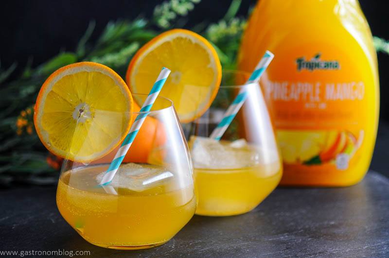 A pair of Tropical Rum Punch cocktails made with Tropicana Pineapple Mango with Lime Premium Juice Drink. Straws and orange wheels