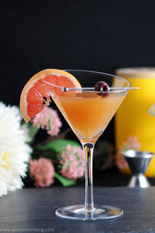The Apple Cider Sidecar in a martini glass with a cherry on a cocktail pick and a slice of grapefruit. A jigger and flowers in the background.