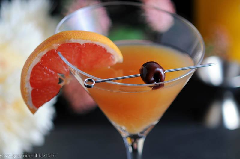 The Apple Cider Sidecar in a martini glass with a cherry on a cocktail pick and a slice of grapefruit. A jigger and flowers in the background.