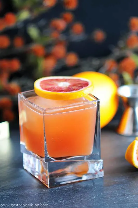 Blood and Bourbon orange cocktail in square rocks glass. Blood orange slice on top of glass. Orange and orange flowers behind glass.