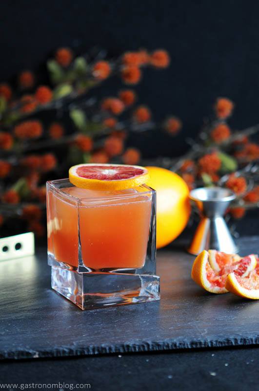 Blood and Bourbon cocktail, orange cocktail in square rocks glass with blood orange slice on top of glass and to the side. Orange, jigger, orange flowers behind glass. 