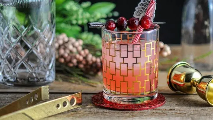 The Red Hound Cocktail made with cranberry, fresh grapefruit, elderflower liqueur and vodka, in a rocks glass and garnished with cranberries and straws, on a wooden table.