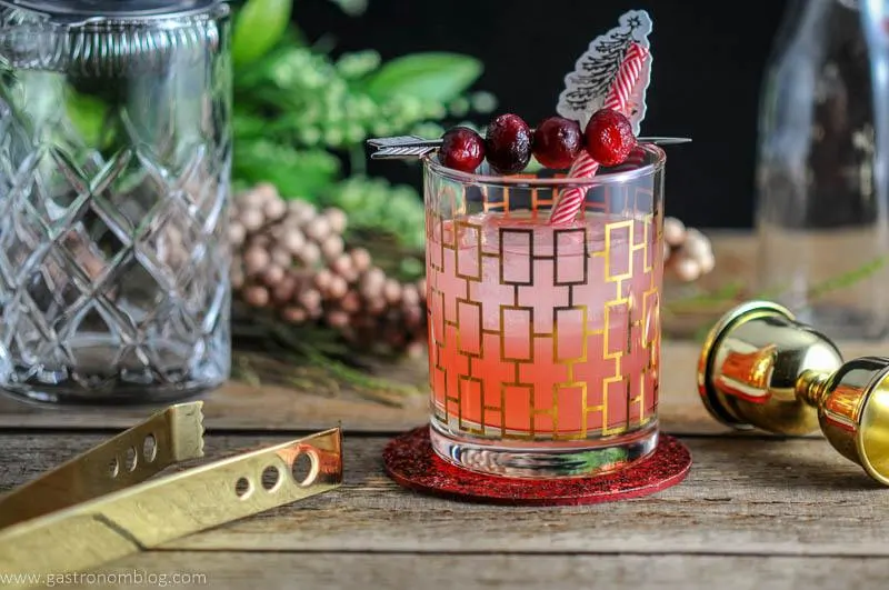 The Red Hound Cocktail in a rocks glass and garnished with cranberries and straws, on a wooden table.